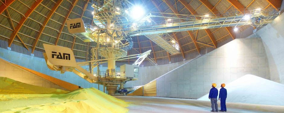 wo workers stand in a large factory hall. In the middle is a large conveyor system, in front of which is a layer of yellow sand-like material.